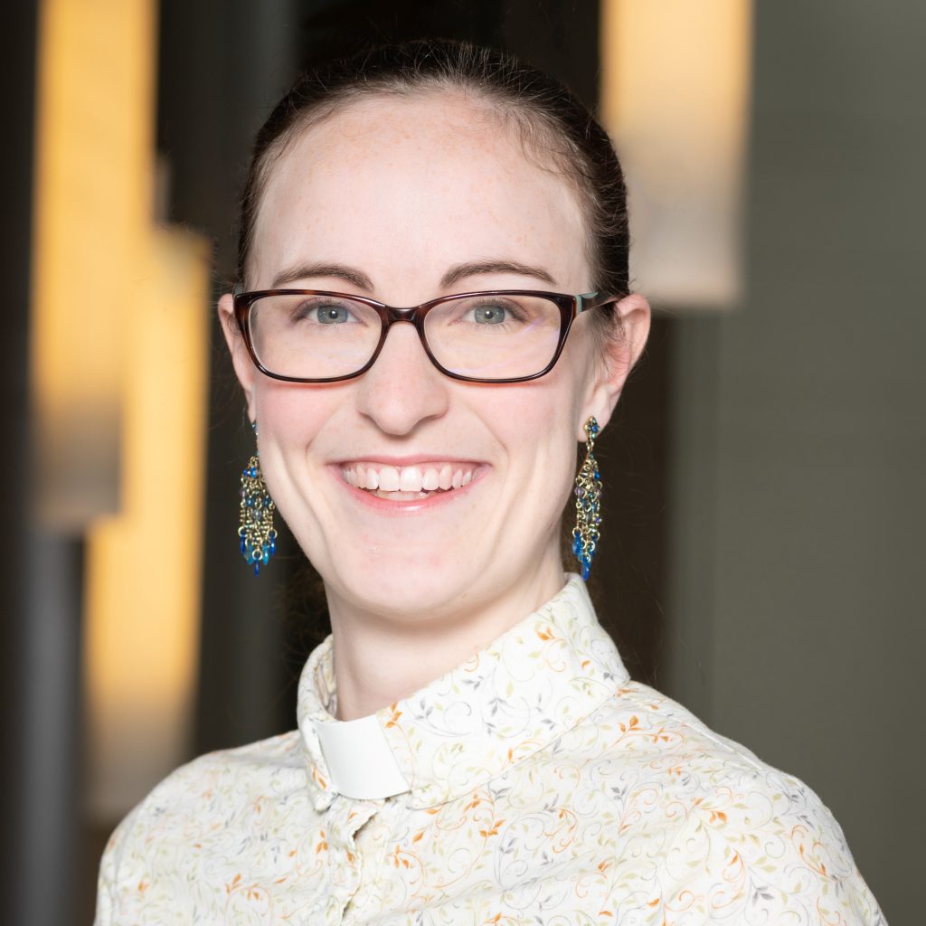 Professional headshot of the Rev. Rachel Erin Stuart. She is a young white woman with dark hair pulled back in a ponytail. She is wearing dark glasses, dangly earrings, and a light floral print clergy shirt.
