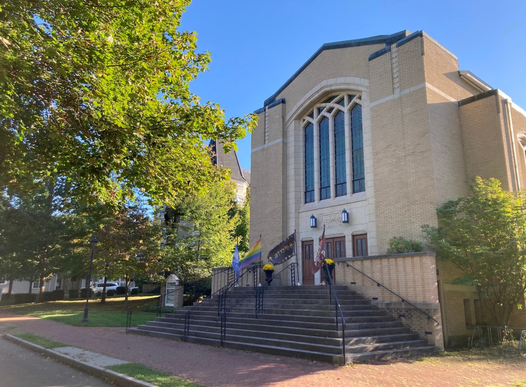 Photo of the exterior of the building in early fall. Trees to the left and right are fading from green toward yellow. Flags emphasizing inclusivity and peace blow gently in the wind upon the front steps.