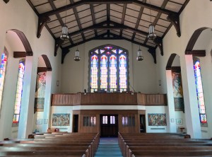Photo of the interior of the sanctuary, facing the narthex. The light suggests early sunset. The balcony is visible, with a large art deco window behind. Several pews are visible, empty at this hour.