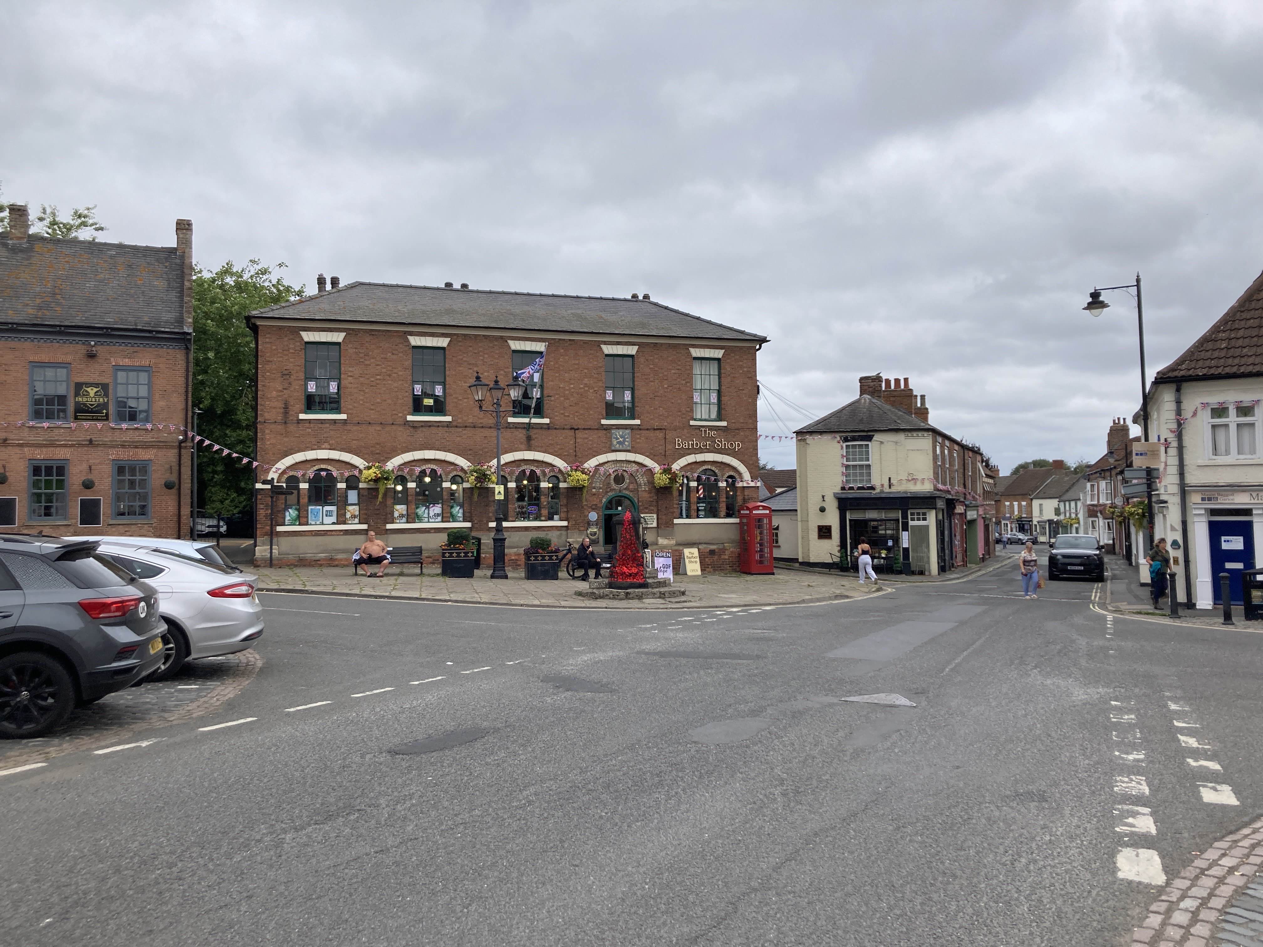 The Epworth Market Cross on a cloudy day. The streets dominate the photo, with a few buildings behind. The stone marker for the crossing is visible in the distance.